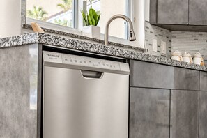 A modern kitchen with a stainless steel dishwasher, granite countertop, and a window with a view of palm trees. - Miami Retreat: Pool, Game Room, Pet-Friendly Space (Miami)