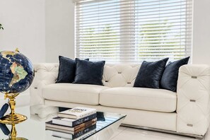 A white leather sofa with dark cushions, a glass coffee table with books, and a globe on a stand. - Miami Retreat: Pool, Game Room, Pet-Friendly Space (Miami)