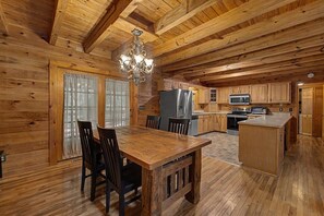 Interior de una cabaña de madera con una mesa de comedor, sillas, una araña y electrodomésticos de cocina.
