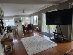 A spacious living room with a large TV on a stand, a dining area with a table and chairs, and a kitchen in the background. - Lakeview Retreat, Deck & Room to Unwind | Get Back 2 Basics Rathmines (Rathmines)
