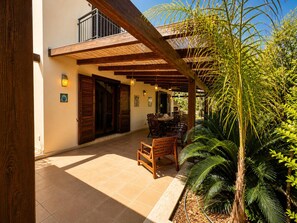 A covered patio area with wooden furniture and a dining table, surrounded by lush greenery and a building with a balcony. - The Secret Garden, villa with private garden (San Vito Lo Capo)