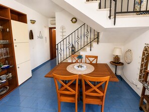 Uma sala de jantar com mesa e cadeiras de madeira, escada com corrimão preto e uma prateleira com copos.
