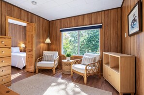 Een kamer met houten lambrisering, een bed, twee rieten stoelen, een tafeltje met een lamp en een groot raam met uitzicht op bomen.