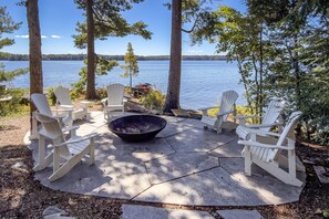 Ulkona on oleskelualue, jossa on Adirondack-tyylisiä tuoleja ja nuotiopaikka, ja sieltä on näkymä järvelle.
