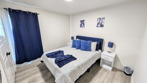 A bedroom with a bed, a nightstand, a lamp, a curtain, and two wall art pieces. - Modern Navy & Black 4BR Near Texas Tech (Lubbock)