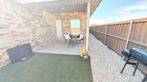 A patio area with a brick wall, two chairs, a small table, a grill, and a covered seating area. - Modern Navy & Black 4BR Near Texas Tech (Lubbock)