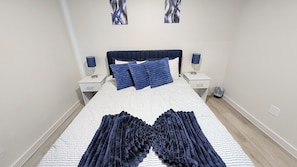 A neatly made bed with a patterned bedspread and blue pillows, flanked by two bedside tables with lamps, in a room with plain walls and a light-colored floor. - Modern Navy & Black 4BR Near Texas Tech (Lubbock)