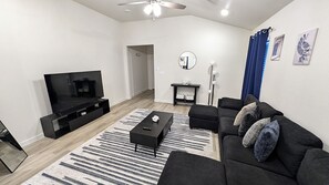 A modern living room with a flat-screen TV, a sofa, a coffee table, and a patterned rug. - Modern Navy & Black 4BR Near Texas Tech (Lubbock)