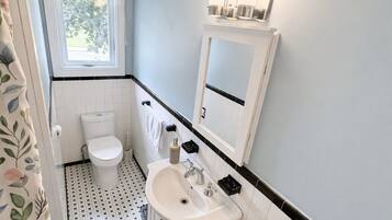 Un baño con inodoro, lavamanos y bañera. Hay una ventana sobre el inodoro y un espejo sobre el lavamanos.