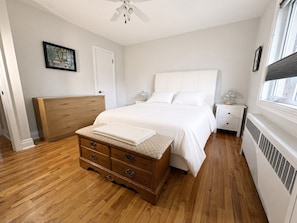 Un dormitorio con cómoda de madera, cama con ropa blanca, mesita de noche, cuadro enmarcado en la pared y ventana con persianas.