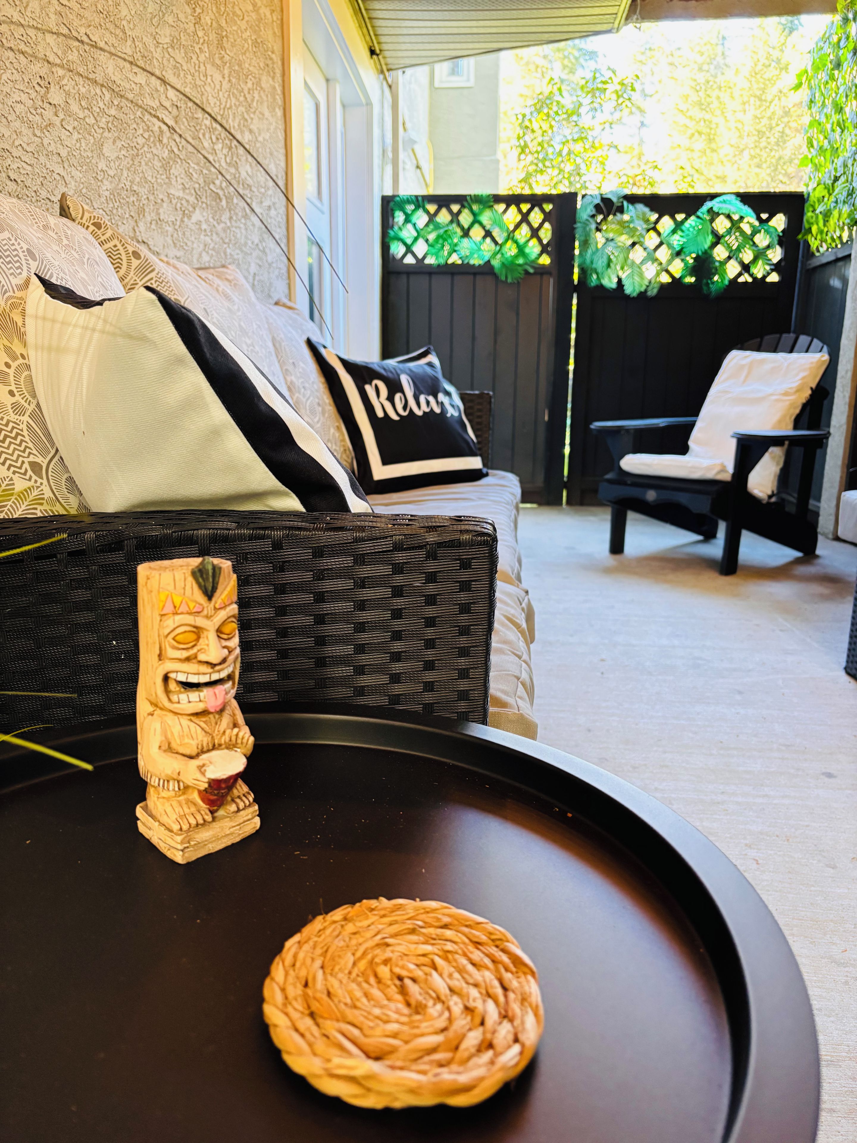 A wicker sofa with a 'Relax' pillow, a woven tray table, and a tiki statue.