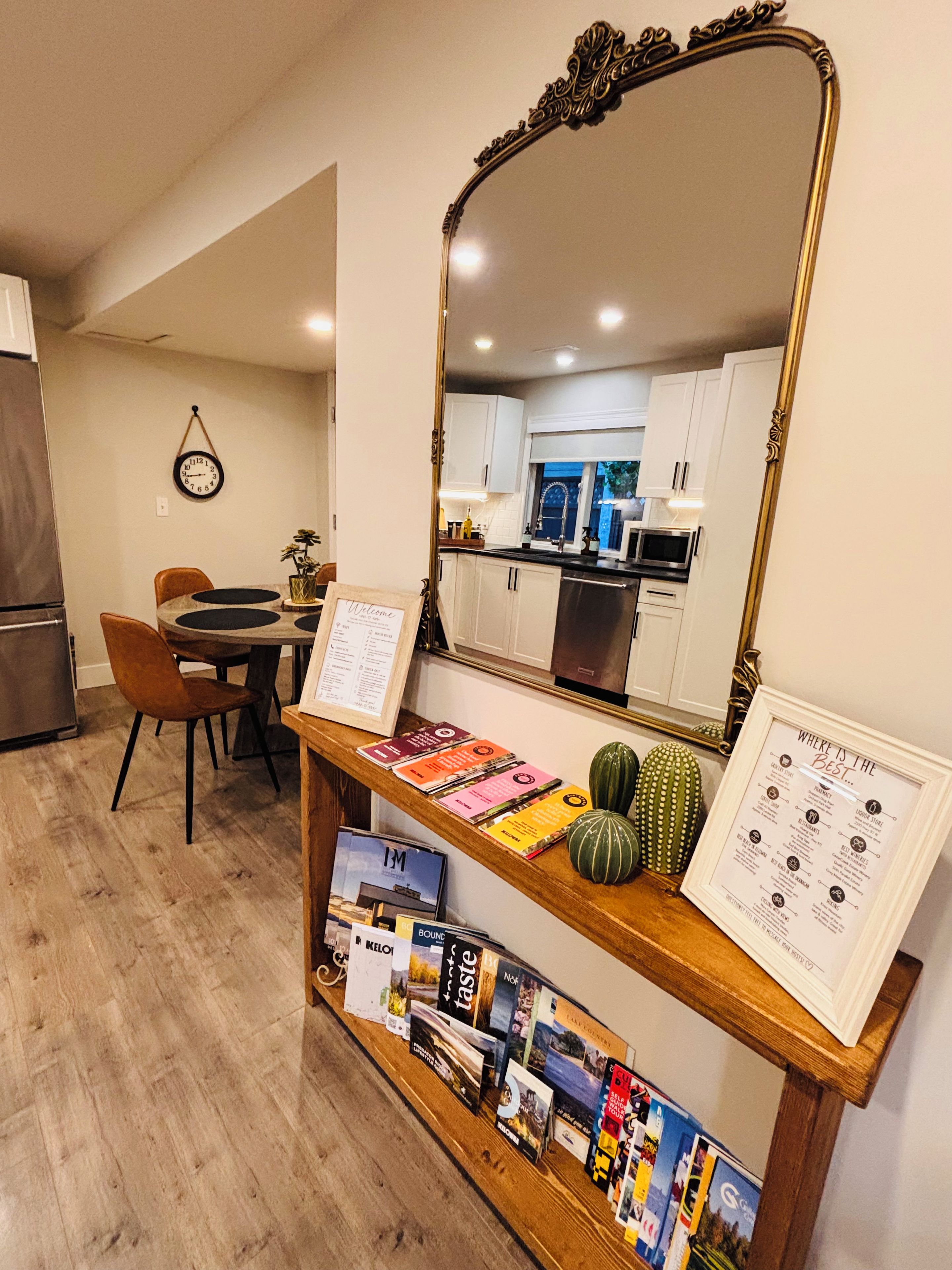 A dining area with a wooden table, chairs, a mirror, and a shelf with books and decorative items.