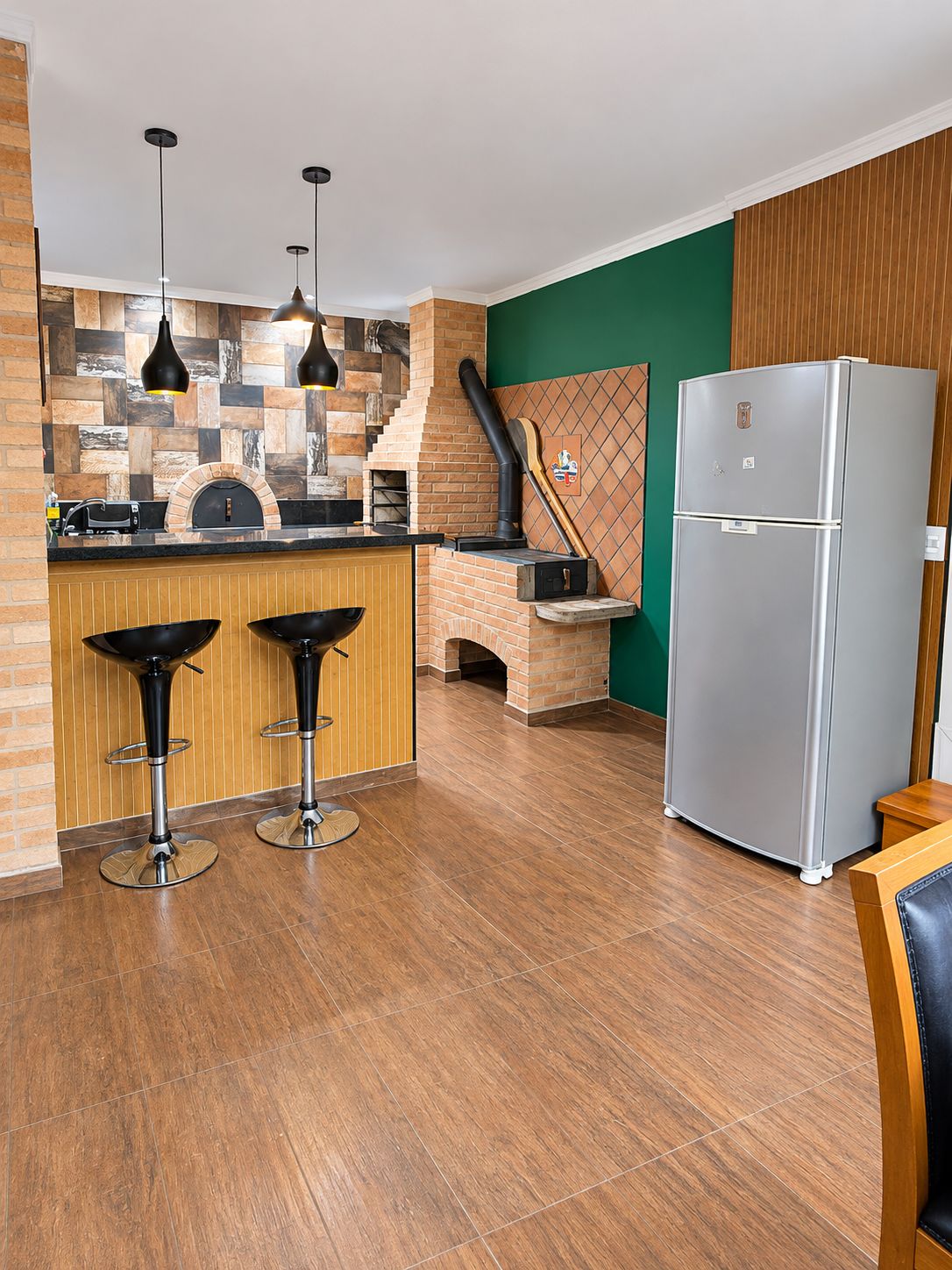 A kitchen with a brick oven, two bar stools, a stainless steel refrigerator, and pendant lighting.
