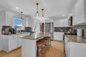A modern kitchen with white cabinetry, a central island with stools, stainless steel appliances, and a view of the outdoors. - 1827 Washington Ave/ Breezy (Seaside Heights)