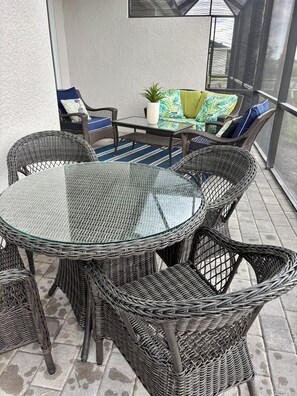 Een glazen ronde tafel omringd door rieten stoelen, een klein tafeltje met een bloempot en een comfortabele bank met kussens.