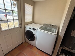 A compact laundry room with a front-loading washer and dryer, a window with blinds, and a view of parked cars outside. - Cowboy Casita~Walk to 11th St. Cowboy Bar (Bandera)