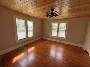 Una habitación con piso de madera, dos ventanas con persianas y un ventilador de techo.