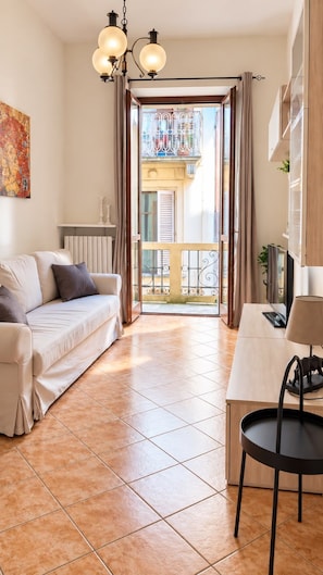 A living room with a sofa, a table, and a chair. There is a balcony with a railing and a view of buildings. - QuickHome Via Italia 43 (Biella)
