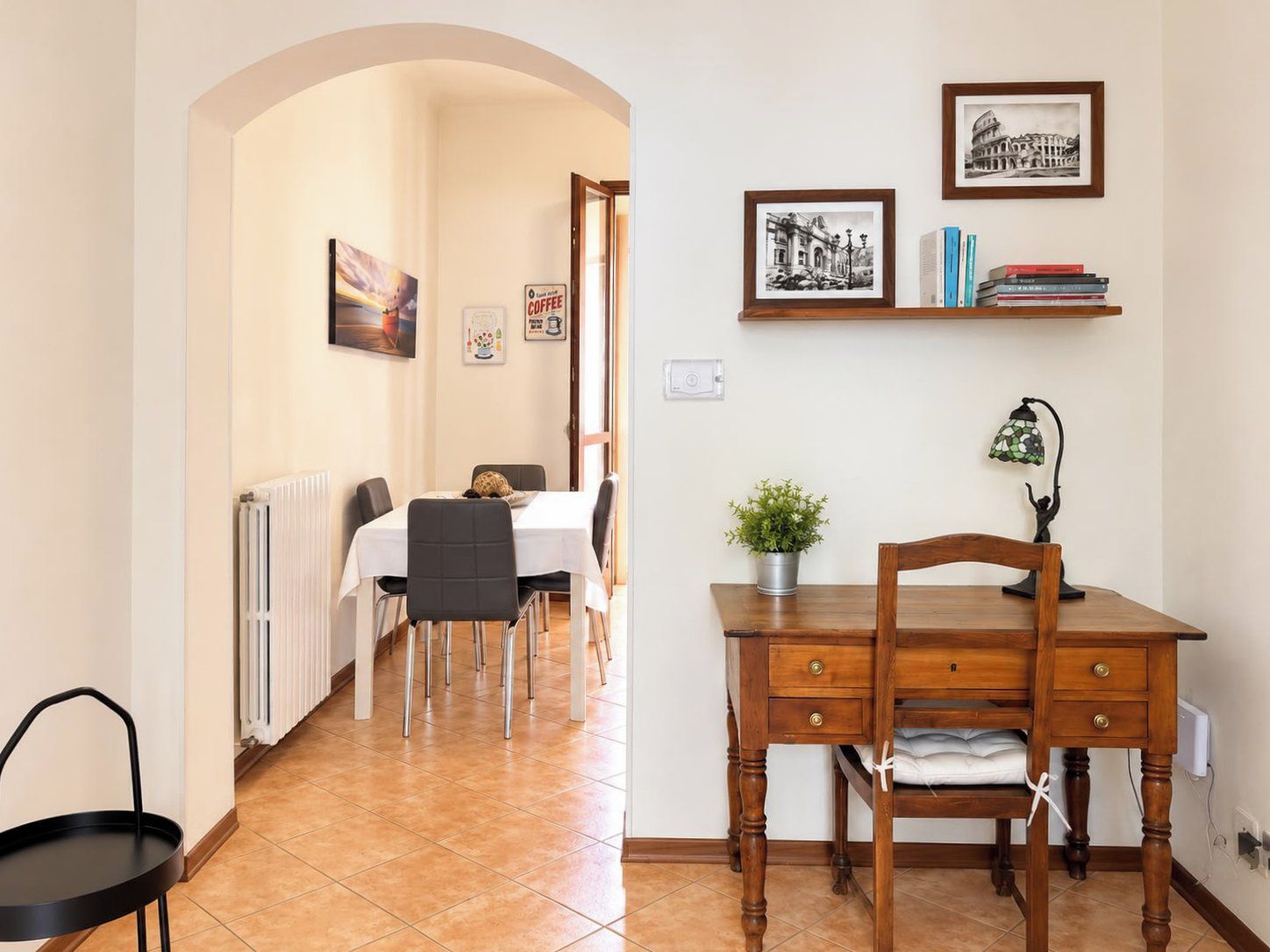 A dining area with a table, chairs, a radiator, a desk with a lamp, and a shelf with books and framed pictures.