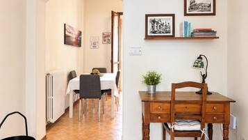 A dining area with a table, chairs, a radiator, a desk with a lamp, and a shelf with books and framed pictures.
