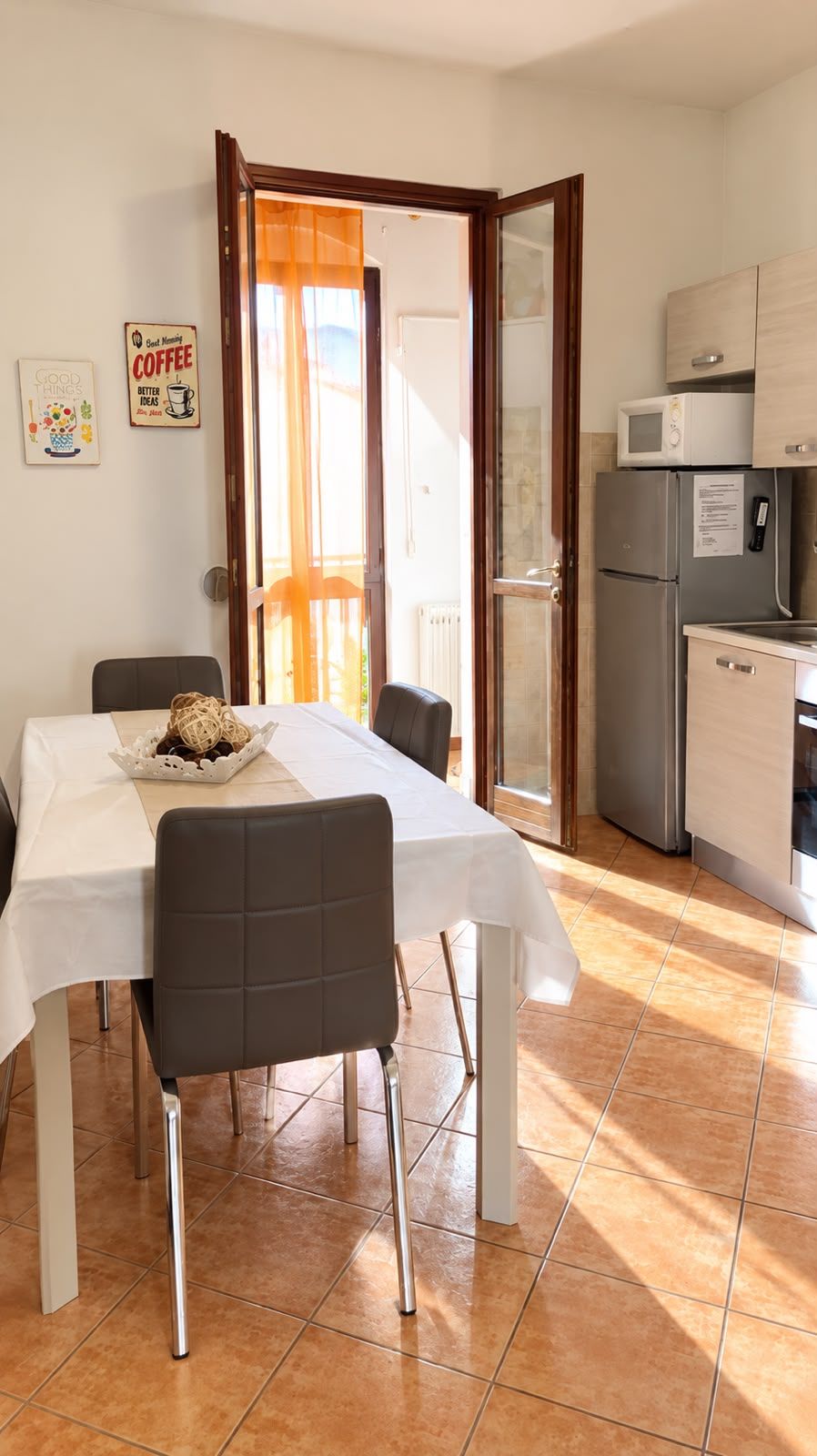 Een eethoek met een tafel en stoelen, een keuken met een koelkast en magnetron, en een deur naar een buitenruimte.