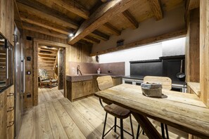 Cozinha rústica com vigas de madeira, área de jantar com mesa e cadeiras de madeira, e vista da cozinha por uma porta.