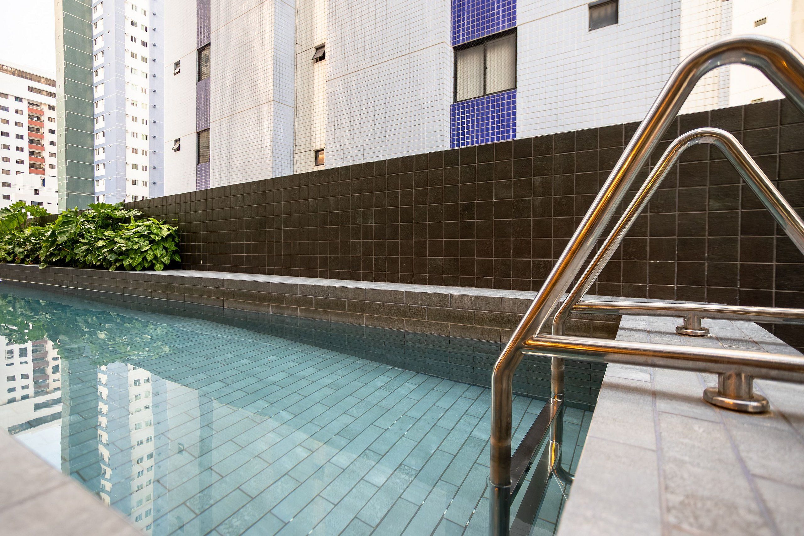Una piscina con una escalera de metal, rodeada por un muro de azulejos y vegetación.