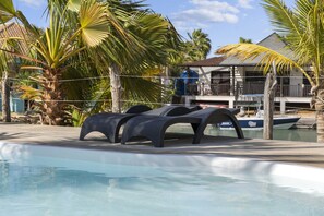 Un espace aménagé au bord de la piscine, avec des chaises longues, des palmiers et un bâtiment en arrière-plan.