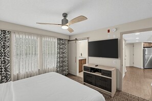 Habitación de hotel con cama, ventilador de techo, televisor y kitchenette.