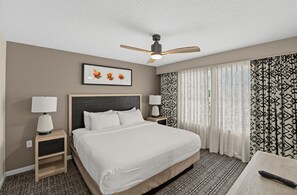 Ein Hotelzimmer mit Bett, Nachttischen, Deckenventilator, einem gerahmten Bild und gemusterten Vorhängen.