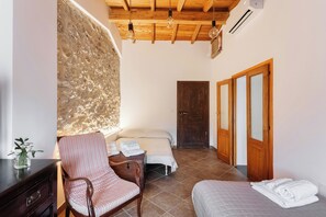 Un dormitori amb paret de pedra, bigues de fusta al sostre, un llit, una cadira i una tauleta de nit.