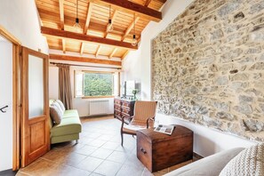 Habitació amb paret de pedra, bigues de sostre de fusta, un sofà verd, un arca de fusta i una finestra amb vista a vegetació.