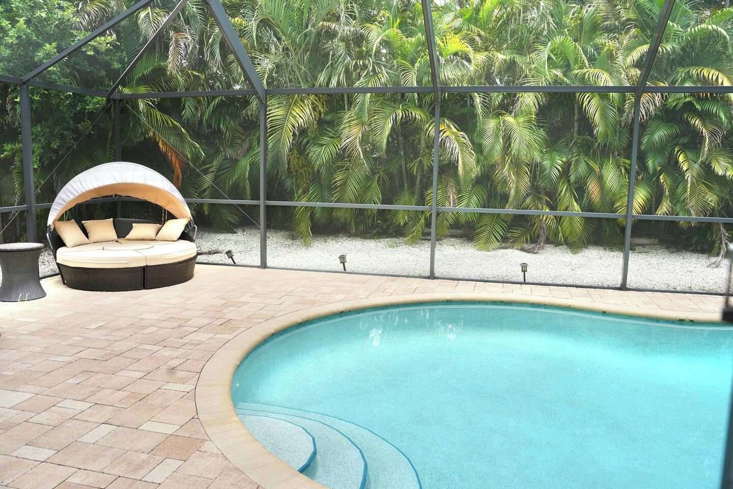 A pool area with a lounge chair and a small table, surrounded by a metal fence and lush greenery.