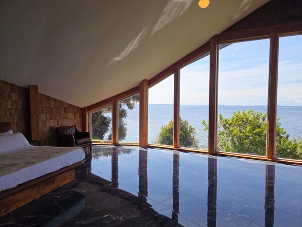Un dormitorio con una ventana grande que da a una masa de agua, una cama y una zona de estar.