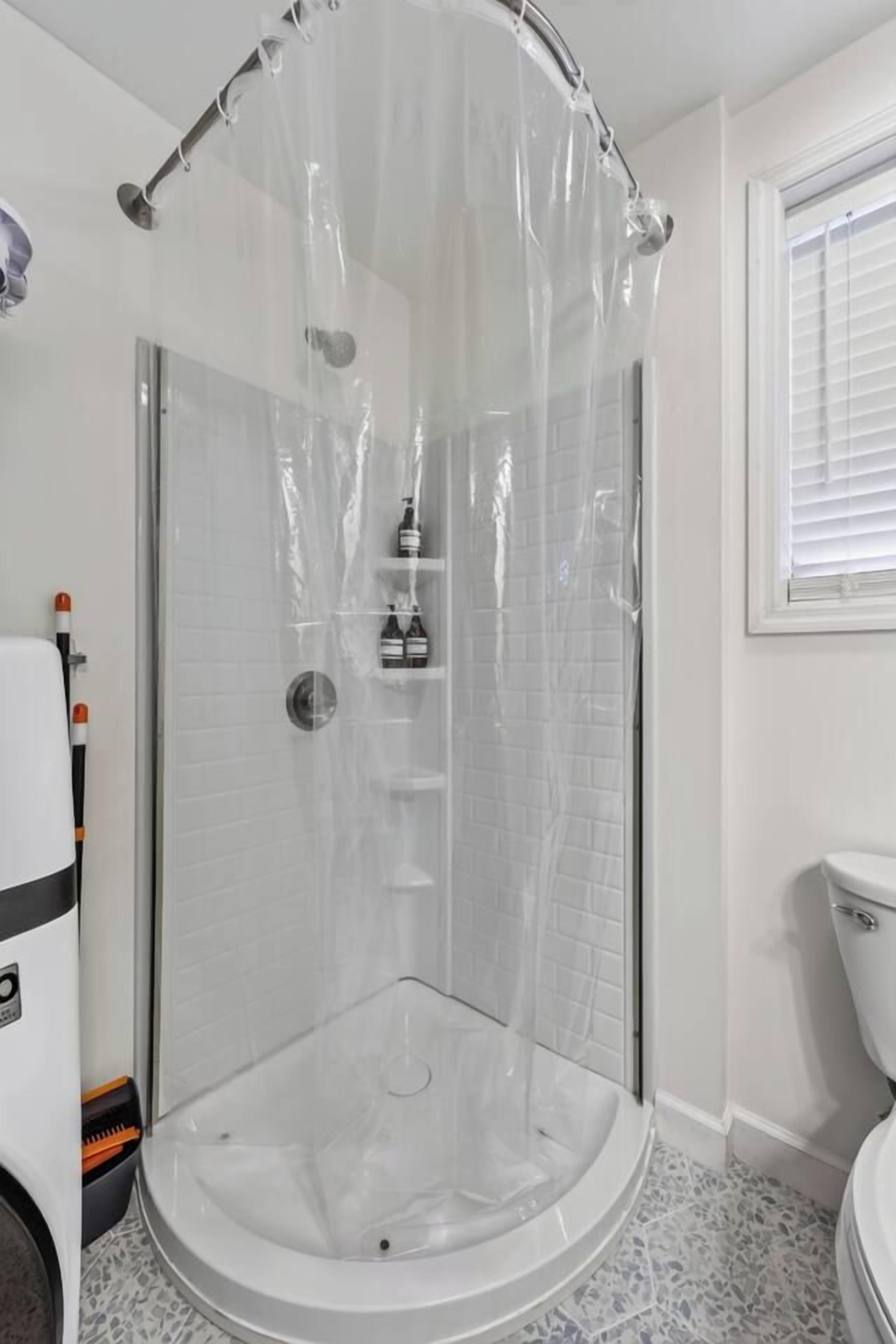 Un box de ducha con cortina de plástico transparente, una pared de azulejos blancos y un inodoro blanco en la habitación contigua.