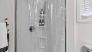Un box de ducha con cortina de plástico transparente, una pared de azulejos blancos y un inodoro blanco en la habitación contigua.