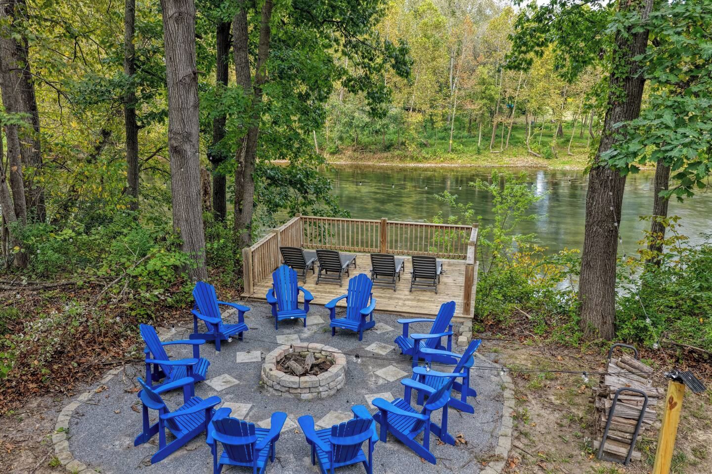 Un espace extérieur aménagé avec des chaises Adirondack bleues disposées autour d’un foyer, avec vue sur un lac.