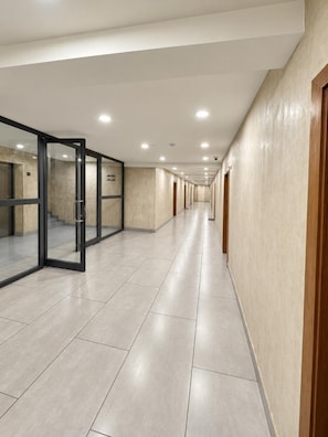Un long couloir étroit, doté d’un sol carrelé, de portes vitrées d’un côté et d’une série de portes de l’autre.