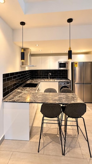 Una cocina moderna con encimera de mármol, muebles negros y blancos, electrodomésticos de acero inoxidable y dos taburetes de bar negros.