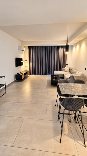 Un salon moderne comprenant un canapé, une table à manger avec des chaises, un téléviseur et une grande fenêtre avec des rideaux.