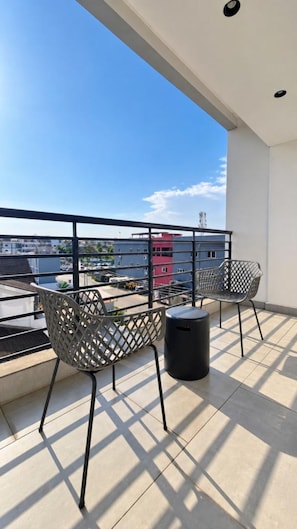 Un balcón con dos sillas y un cubo de basura, con vista a un paisaje urbano con edificios y cielo despejado.