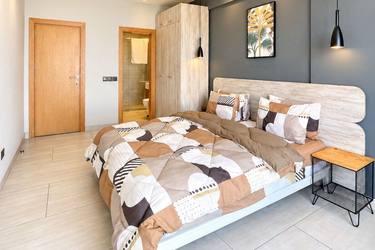 Une chambre à coucher moderne, avec un grand lit, une table de chevet, une armoire et une salle de bain visible à travers une porte ouverte.