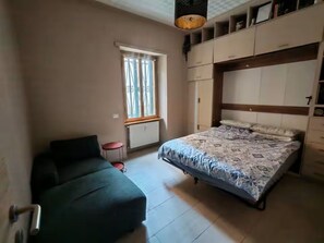 A bedroom with a bed, a sofa, a window, and built-in storage. - New Listing (Roma)