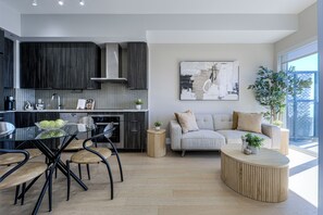A modern kitchen with dark cabinetry, a dining area with a glass table and chairs, a grey sofa with cushions, and a large abstract painting on the wall. - Waterfront Modern 1-Bedroom Condo With Parking (Toronto)