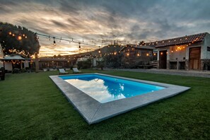 Ein Swimmingpool, umgeben von einer gepflegten Rasenfläche, mit Lichterketten und rustikalen Gebäuden im Hintergrund.