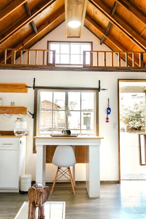 A room with a lofted wooden ceiling, a desk with a chair, a window with a view, and a hanging ornament. - Long Stay Cabin 2 - by Hacienda Cuatro Milpas (Ensenada)