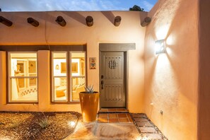 A building entrance with a large potted plant, a door with a security lock, and a window overlooking the interior. - Casa Altura - Hot Tub Desert Retreat - Pet Friendly (Las Cruces)