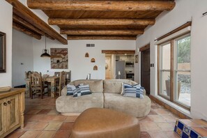 A cozy living room with a wooden ceiling, a sofa with cushions, a dining table with chairs, and a view through a large window. - Casa Altura - Hot Tub Desert Retreat - Pet Friendly (Las Cruces)