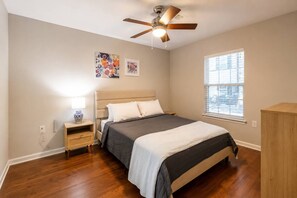 Quarto com cama, mesa de cabeceira, ventoinha no teto e janela.