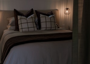 Cama bem arrumada com cabeceira escura, roupa de cama branca e manta marrom.
